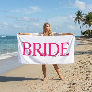 May include: White beach towel featuring the word "BRIDE" in vibrant pink, with "Mrs. Diana" in cursive. The towel is displayed on a sandy beach, with the sea and palm trees visible in the background.