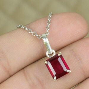 Puede incluir: Un collar de plata con un colgante rectangular de piedra preciosa rojo oscuro. El colgante está engastado en un marco plateado y cuelga de una delicada cadena. La piedra preciosa tiene una superficie pulida y reflectante.