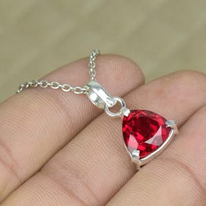 Puede incluir: Un collar de plata con un colgante triangular facetado de piedra preciosa rojo intenso. El colgante está engastado en un bisel de plata y cuelga de una delicada cadena de plata. La piedra preciosa tiene un corte brillante.