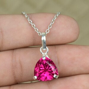 Puede incluir: Un collar de plata con un colgante triangular de piedra preciosa fucsia. El colgante cuelga de una delicada cadena. La gema facetada brilla. Este collar es una joya.