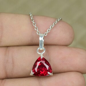 Può includere: Una collana d'argento con un pendente triangolare sfaccettato in pietra preziosa rosso intenso. Il pendente è incastonato in una lunetta d'argento e pende da una delicata catena d'argento. La gemma ha una superficie brillante e riflettente.
