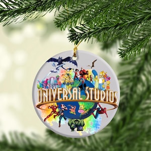 May include: A round, white Christmas ornament featuring the Universal Studios logo with various cartoon characters and superheroes. The ornament is hanging from a gold string and is set against a blurred green background.