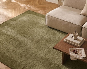May include: A rectangular area rug in a solid olive green color featuring a distinct border design. The rug has a textured, woven surface and is positioned on a light-colored wooden floor with a herringbone pattern. The text The Classic Rugs is visible.