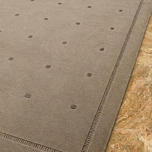May include: A rectangular area rug in a solid taupe color featuring a repeating pattern of small, dark square accents across the surface. The rug is finished with a wide, textured border that frames the central field of the design.