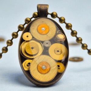 Puede incluir: Collar con colgante ovalado con una colección de engranajes de reloj dorados encapsulados en resina transparente. El colgante cuelga de una cadena de bolas de color bronce a juego. Los engranajes tienen detalles en naranja y amarillo.