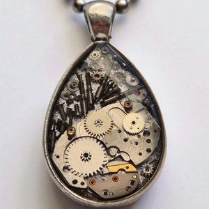 Puede incluir: Un collar con colgante plateado con un colgante en forma de lágrima lleno de intrincados engranajes y componentes de reloj. Los engranajes son de varios tamaños y formas, creando una estética steampunk. El collar tiene una cadena de bolas.
