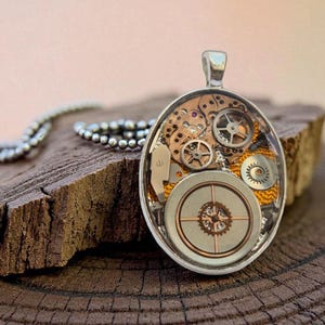 Puede incluir: Colgante ovalado con diseño steampunk. El colgante plateado presenta engranajes y elementos de relojería visibles, encapsulados en resina transparente. El collar descansa sobre un trozo de madera marrón oscuro, con una cadena plateada visible.
