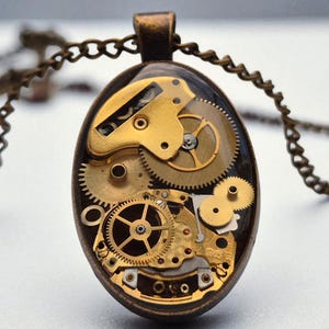 Puede incluir: Collar con colgante ovalado con diseño steampunk. El colgante muestra intrincados engranajes dorados y componentes mecánicos encapsulados en resina transparente. El collar tiene una cadena de color bronce.