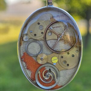 Puede incluir: Collar con colgante ovalado que presenta engranajes y componentes de reloj encapsulados en resina transparente. El colgante tiene un marco de metal plateado y cuelga de una cadena de bolas. El fondo es verde borroso.