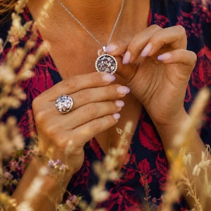 Puede incluir: Un collar de tono plateado con un colgante circular que presenta un centro oscuro texturizado y multifacético rodeado por un borde de pequeñas piedras claras, junto con un anillo circular a juego con un diseño oscuro y facetado similar.