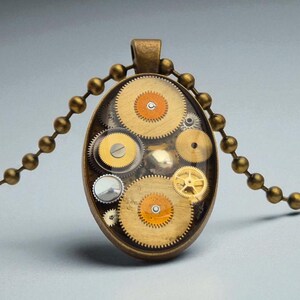 Puede incluir: Collar con colgante ovalado con una colección de engranajes de latón encapsulados en resina transparente. El colgante cuelga de una cadena de bolas de latón a juego. Los engranajes tienen detalles en naranja y amarillo, creando una estética steampunk.