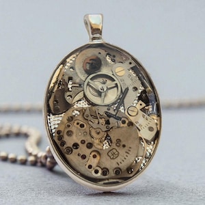 Puede incluir: Collar con colgante ovalado que muestra los intrincados mecanismos de un reloj. El colgante plateado revela engranajes, muelles y otros componentes mecánicos encapsulados en resina transparente. El collar tiene una fina cadena.