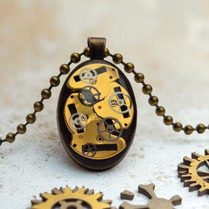 Puede incluir: Collar con colgante ovalado con un diseño steampunk, que muestra engranajes y ruedas dentadas doradas dentro de un marco marrón oscuro. El colgante cuelga de una cadena de bolas de color bronce. Varios componentes sueltos en forma de engranaje están dispersos alrededor del colgante.