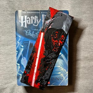 May include: A bookmark featuring Darth Maul from Star Wars, with a red lightsaber design. The bookmark is placed in a book titled "Harry Potter and the Order of the Phoenix". The bookmark has a black background with red accents.