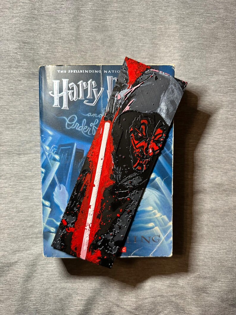 May include: A bookmark featuring Darth Maul from Star Wars, with a red lightsaber design. The bookmark is placed in a book titled "Harry Potter and the Order of the Phoenix". The bookmark has a black background with red accents.