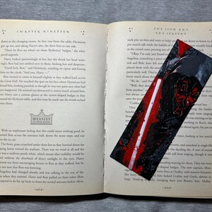 May include: A handmade bookmark with a black and red abstract design, resting on an open book. The bookmark features a textured, painted appearance. The book pages are aged, with text visible.