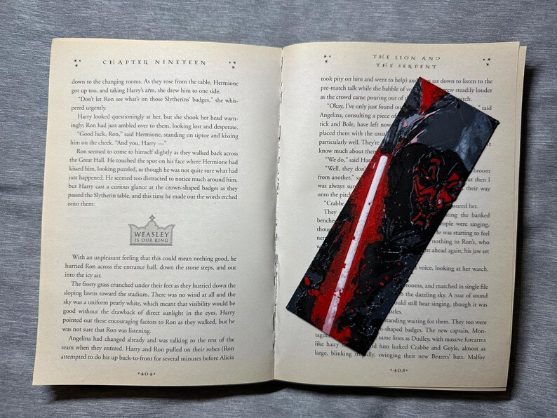May include: A handmade bookmark with a black and red abstract design, resting on an open book. The bookmark features a textured, painted appearance. The book pages are aged, with text visible.