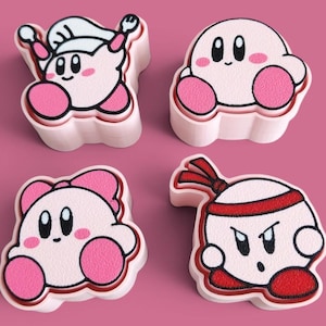 May include: Four light pink, three-dimensional cookie cutters featuring the character Kirby in various forms. One wears a chef hat, one is a standard round shape, one has a pink bow, and one wears a red headband with a determined facial expression.