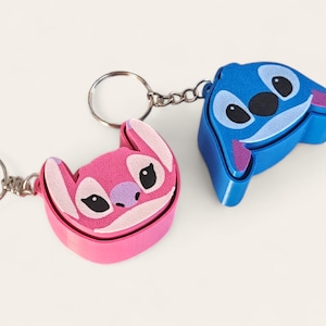 May include: Two character keyrings, one pink and one blue. The pink keyring features a character with a light purple nose and ears. The blue keyring has a white face and pink ears. Both have black eyes.