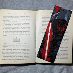 May include: A handmade bookmark featuring a Star Wars Darth Maul design. The bookmark is rectangular with a red, black, and white color scheme. It is placed inside an open book with text visible on the pages.