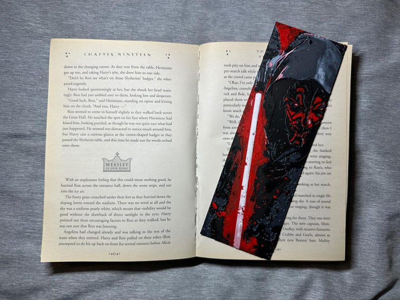 May include: A handmade bookmark featuring a Star Wars Darth Maul design. The bookmark is rectangular with a red, black, and white color scheme. It is placed inside an open book with text visible on the pages.