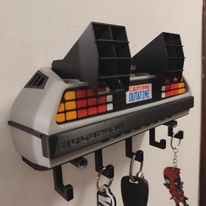 May include: A wall-mounted key holder designed to resemble the rear of a silver sports car. It features black vents, red and orange taillights, and a California OUTATIME license plate. Five black hooks extend from the bottom to hold various keychains.
