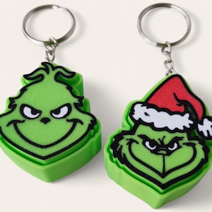 May include: Two green keychains featuring the face of a cartoon character with a mischievous expression. One keychain shows the character alone, while the other depicts the character wearing a red and white Santa hat with a silver metal ring.