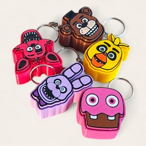 May include: A selection of character keyrings in various colours. The keyrings depict a bear, a chicken, a rabbit, a cupcake, and a fox. Each keyring has a silver metal ring attached.