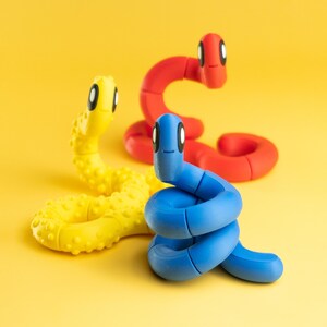 May include: Three colourful, segmented worm toys in yellow, red, and blue, each with a smiling face and black and white eyes. The yellow worm has a textured surface. The worms are arranged on a bright yellow background.