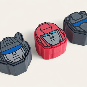 May include: Three geometric boxes featuring robot face designs. One is red, one is grey with blue accents, and the other is dark grey with blue accents. These boxes are likely for storage or display.