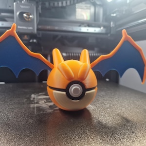 May include: A 3D-printed orange and blue Charizard-themed Pokeball. The wings are blue with orange trim, and the ball has a white and black centre. The design is a stylized representation of the popular character.