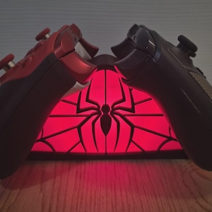 May include: A red and black gaming controller charging stand featuring a glowing red spider emblem and web pattern. The stand is positioned between two wireless gaming controllers, one in red and one in black, resting on the charging base.