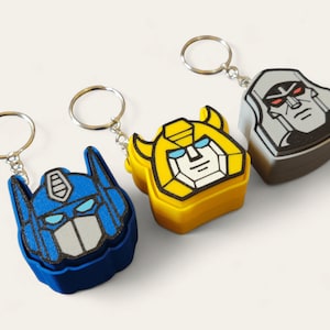 May include: Three character-shaped keyrings. One is blue with a white face, one is yellow with a white face, and the last is grey with a black face. Each keyring has a silver ring attached.