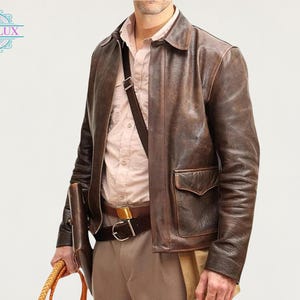 Könnte beinhalten: Braune Lederjacke mit zwei Vordertaschen und einem Kragen. Die Jacke wird über einem hellrosa Button-Down-Hemd getragen. Ein brauner Ledergürtel und eine Umhängetasche sind ebenfalls zu sehen. Das Bild enthält den Text "OBSILUX".