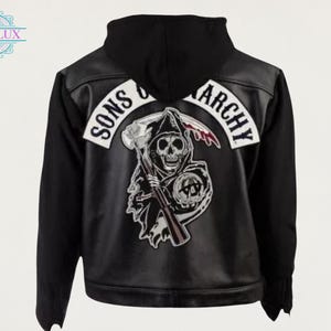 Puede incluir: Chaqueta negra con capucha y las palabras "SONS OF ANARCHY" en la espalda. La chaqueta presenta un gran parche con una parca sosteniendo una guadaña y una calavera. La chaqueta está hecha de una combinación de cuero negro y tela.