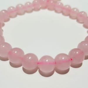 Könnte beinhalten: Ein Armband aus runden, hellrosa Rosenquarzperlen. Die Perlen sind mit einem rosa Faden aufgefädelt. Das Armband wird auf einer weißen Oberfläche gezeigt. Die Perlen haben einen Durchmesser von ca. 1 cm.