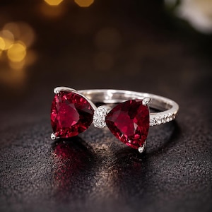 May include: A silver ring featuring two deep red, trillion-cut gemstones arranged to resemble a bow. The center of the bow is accented with small, clear pavé stones, and the ring band is also set with a row of small, clear, sparkling stones.