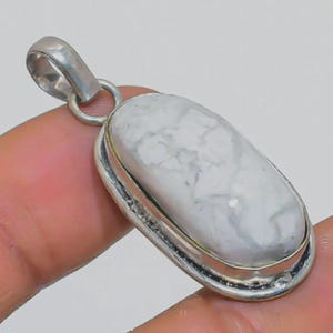 May include: A silver pendant with an oval, white stone featuring gray marbling. The stone is set in a silver bezel and has a silver loop for a chain. The pendant is held by a hand.
