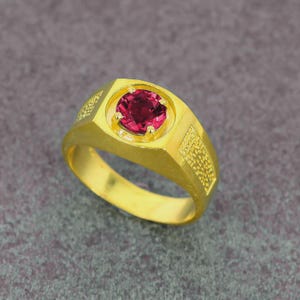 Puede incluir: Un anillo dorado con una gema roja grande y redonda engastada en un bisel cuadrado. El anillo tiene un diseño texturizado en los lados y un acabado pulido. El anillo es de estilo sello clásico.