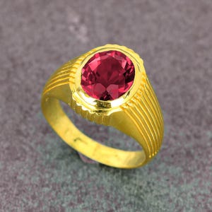 May include: A gold ring featuring a large, oval-cut, deep red gemstone. The ring has a textured, ridged band and a decorative bezel around the stone. The ring is set against a gray background.