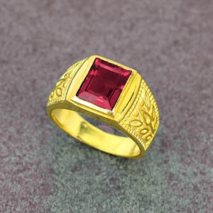 Könnte beinhalten: Ein goldener Ring mit einem rechteckigen, tiefroten Edelstein. Der Ring hat ein verziertes Design am Band mit poliertem Finish. Der Edelstein ist in einer quadratischen Fassung gefasst, was einen klassischen Look ergibt.