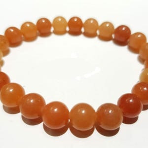 Pode incluir: Uma pulseira com uma sequência de contas redondas de cor laranja. As contas têm uma superfície lisa e polida e variam em tonalidade, de um laranja claro a um laranja avermelhado mais profundo. A pulseira é exibida sobre um fundo branco.