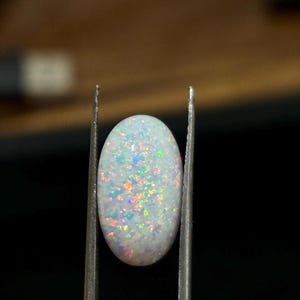 May include: An oval opal gemstone, held by tweezers. The opal has a milky white base colour with iridescent flashes of blue, green, and orange. The gemstone is the focal point against a dark background.