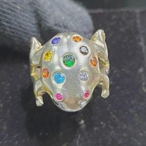 May include: Silver ring in a frog design, embellished with a variety of coloured gemstones. The ring's body is studded with gems in shades of blue, green, yellow, and red. The frog's legs and head are clearly defined, with a textured finish.