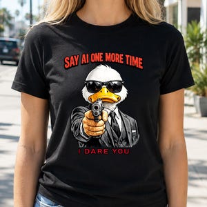 May include: Black t-shirt featuring a cartoon duck wearing sunglasses and a suit, holding a gun. The text "SAY AI ONE MORE TIME" is above the duck, and "I DARE YOU" is below. The duck has a white head, yellow beak, and is pointing the gun forward.