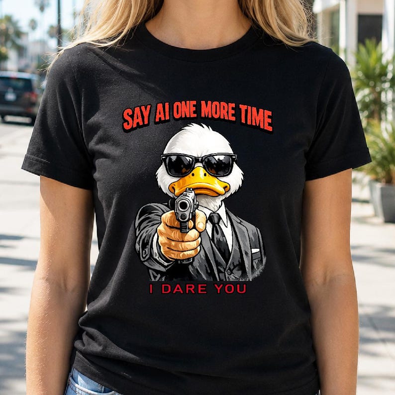 May include: Black t-shirt featuring a cartoon duck wearing sunglasses and a suit, holding a gun. The text "SAY AI ONE MORE TIME" is above the duck, and "I DARE YOU" is below. The duck has a white head, yellow beak, and is pointing the gun forward.