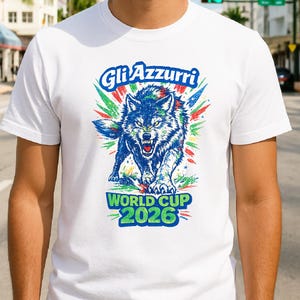 Puede incluir: Camiseta blanca con un lobo azul rugiente. El texto "Gli Azzurri" está arriba y "World Cup 2026" abajo. El diseño incluye toques de verde y rojo, con un aspecto desgastado y vintage.