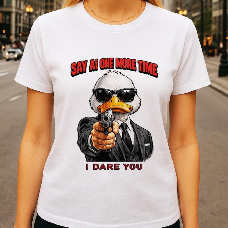 May include: White t-shirt featuring a cartoon duck wearing sunglasses and a suit, holding a gun. The text "SAY AI ONE MORE TIME" is above the duck, and "I DARE YOU" is below.