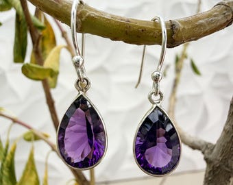 May include: A pair of drop earrings featuring faceted, pear-shaped purple gemstones set in polished silver-toned metal. Each earring hangs from a simple silver-toned hook wire, showcasing the deep violet colour and geometric cut of the stones.