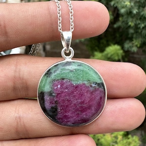 May include: A round pendant necklace featuring a polished ruby zoisite gemstone with vibrant magenta, forest green, and black mineral inclusions. The stone is set in a simple silver-toned metal bezel and hangs from a matching silver-toned chain.